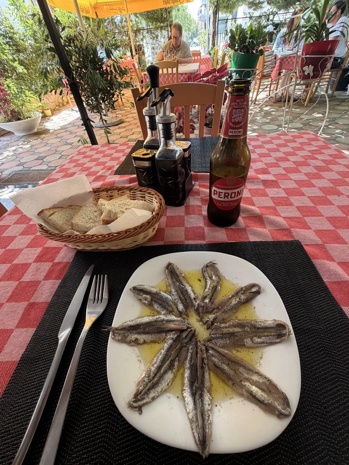 Un piatto di alici fritte disposte a stella su una tovaglia a quadretti rossi a Valona, con pane e una birra.