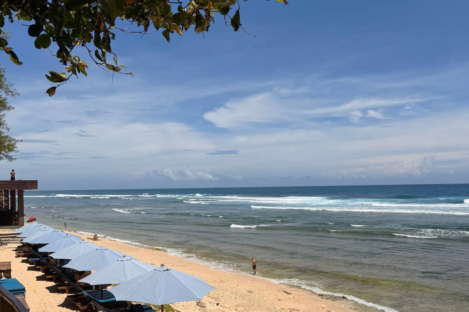 A Bali beach club with blue umbrellas on golden sand and waves rolling in near Canggu.
