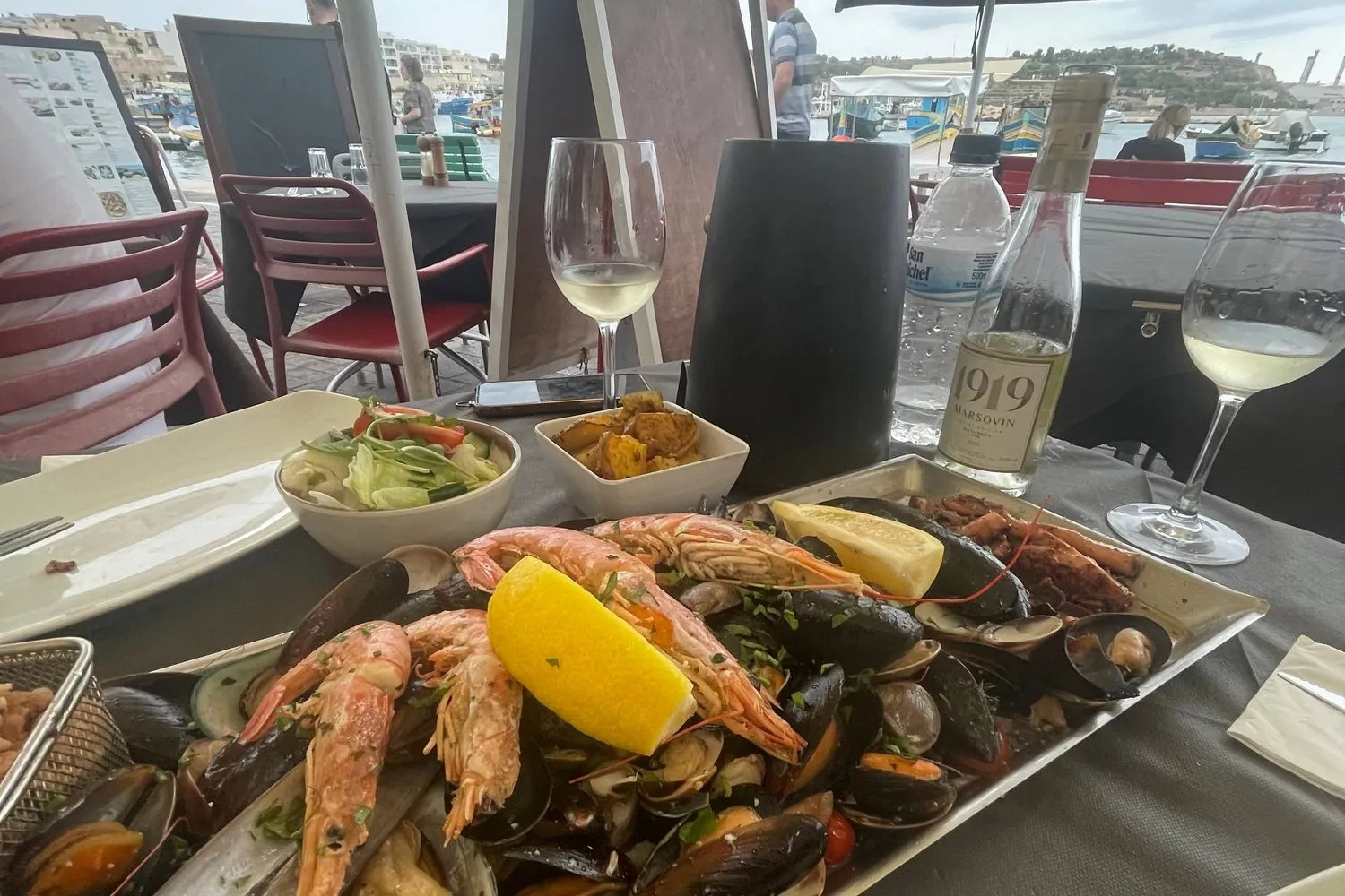 A harbourside seafood platter with mussels and prawns, boats and a Maltese town in the background.
