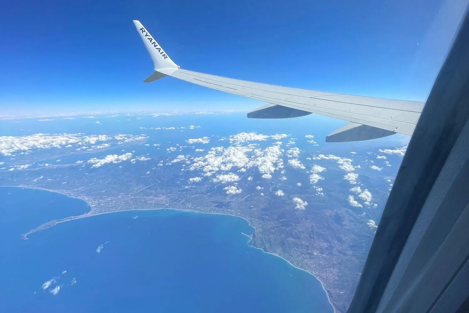 A Ryanair wing over a blue coastline, seen from the plane window.