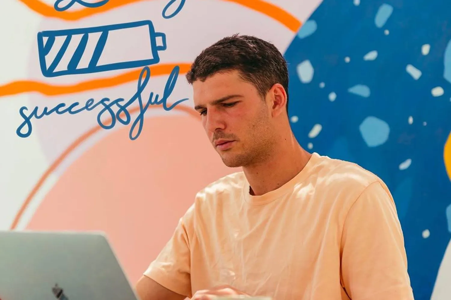 A man working on a laptop with a coffee in front of a colourful coworking mural that reads "Loading successful".